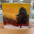 Encaustic painting of snowy forest with pine tree and leafless bushes, orange and yellow sunset in background